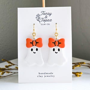 May include: White ghost-shaped earrings with orange bow accents and gold-colored hooks. The earrings have black eyes and a smiling mouth. The card behind the earrings reads "Tansy & Topaz Clay Co." and "handmade clay jewelry."