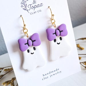 May include: A pair of handmade clay ghost earrings with purple bow accents and gold-colored ear wires. The ghosts have smiling faces. The text "& Topaz Clay Co." and "handmade clay jewelry" are visible.