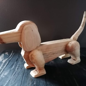 Könnte beinhalten: Eine Holzskulptur eines Dackelhundes. Der Hund ist aus hellem Holz gefertigt, mit sichtbarer Maserung und natürlicher Oberfläche. Er hat einen langen Körper, kurze Beine und einen gebogenen Schwanz. Die Skulptur wird auf einer dunklen Oberfläche präsentiert.