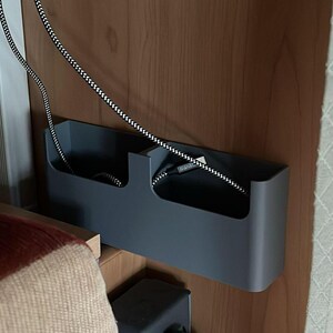 K&ouml;nnte beinhalten: Ein grauer Kabelorganizer mit zwei F&auml;chern, montiert an einer Holzoberfl&auml;che. Ein schwarz-wei&szlig; gestreiftes Kabel h&auml;ngt von der Oberseite des Organizers.