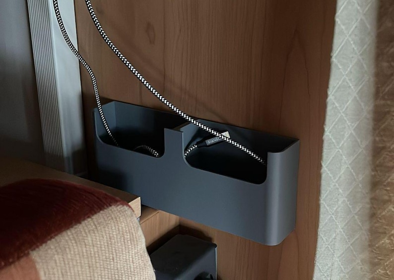 K&ouml;nnte beinhalten: Ein grauer Kabelorganizer mit zwei F&auml;chern, montiert an einer Holzoberfl&auml;che. Ein schwarz-wei&szlig; gestreiftes Kabel h&auml;ngt von der Oberseite des Organizers.