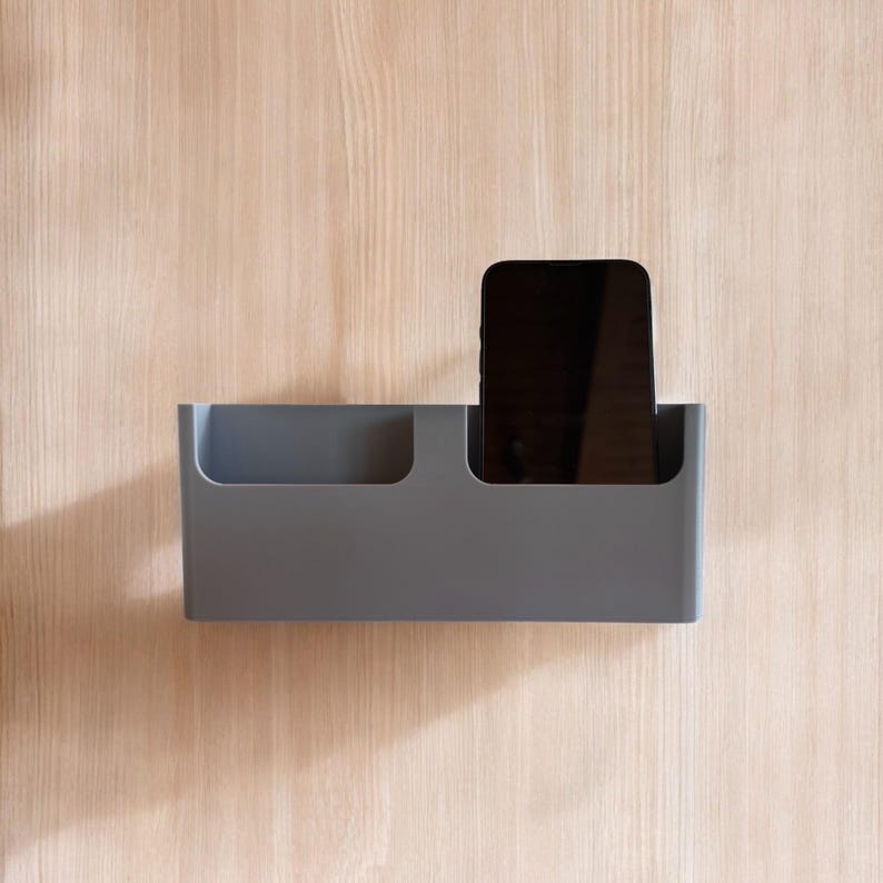 K&ouml;nnte beinhalten: Ein grauer, wandmontierter Organizer mit zwei F&auml;chern, von denen eines ein schwarzes Smartphone enth&auml;lt.