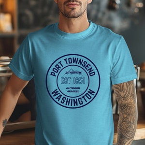A man in a coffee shop wears a heather aqua bella canvas 3001 with a circle graphic reading port townsend washington authentic outdoor apparel.