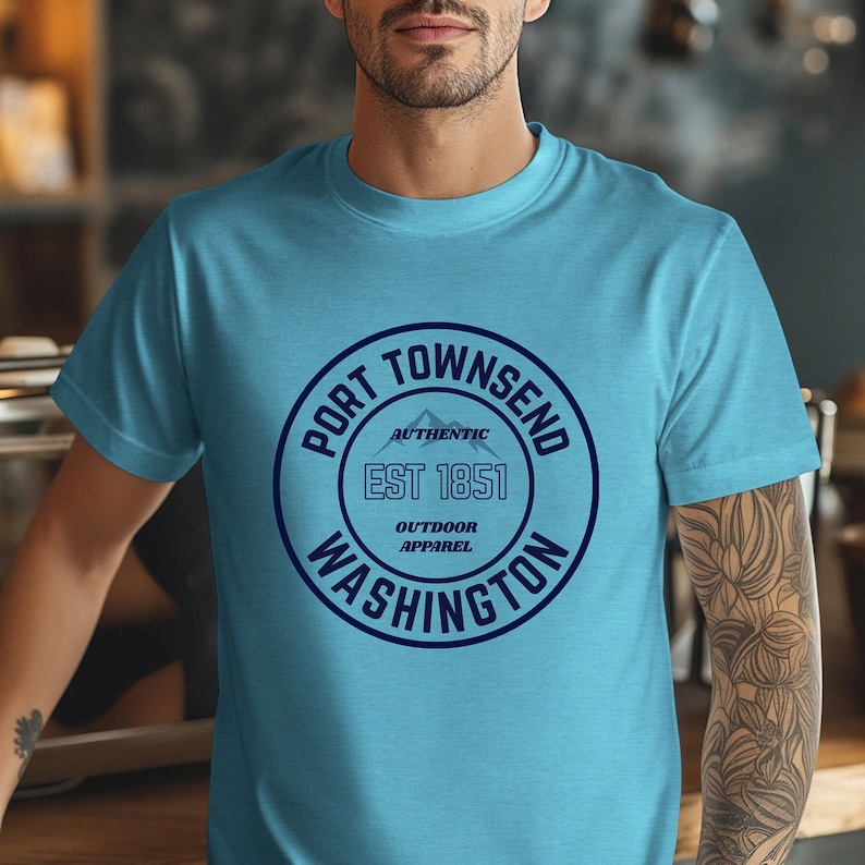 A man in a coffee shop wears a heather aqua bella canvas 3001 with a circle graphic reading port townsend washington authentic outdoor apparel.