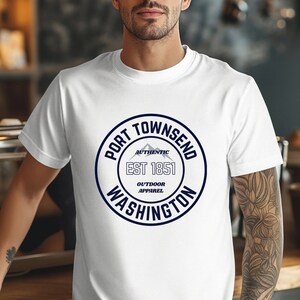 A man in a coffee shop wears a white bella canvas 3001 with a circle graphic reading port townsend washington authentic outdoor apparel.
