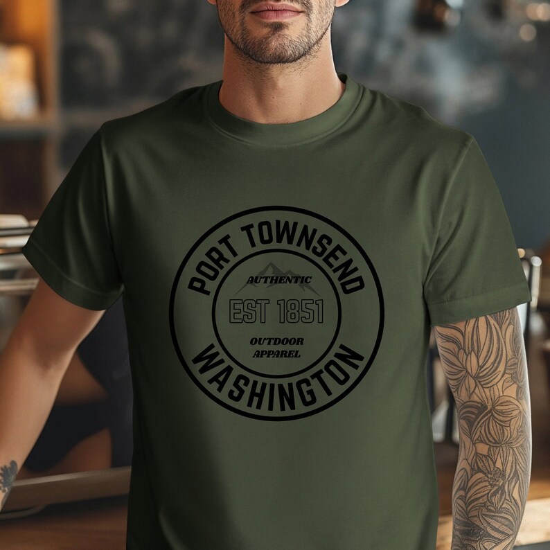 A man in a coffee shop wears a military green bella canvas 3001 with a circle graphic reading port townsend washington authentic outdoor apparel.