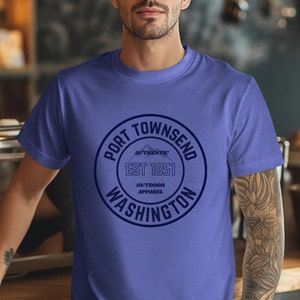 A man in a coffee shop wears a heather true royal aqua bella canvas 3001 with a circle graphic reading port townsend washington authentic outdoor apparel.