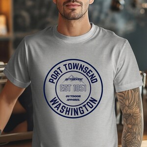 A man in a coffee shop wears an athletic heather aqua bella canvas 3001 with a circle graphic reading port townsend washington authentic outdoor apparel.