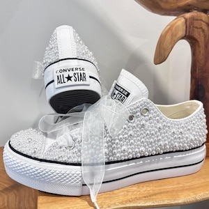 May include: White Converse All Star sneakers embellished with faux pearls. The platform soles are white with a black stripe. The laces are white ribbons. The heel features the Converse All Star logo.