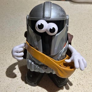 Könnte beinhalten: Eine Spielzeugfigur, die einem Charakter aus einem Science-Fiction-Franchise ähnelt. Die Figur hat einen silbernen Helm, ein schwarzes Visier und große Wackelaugen. Sie trägt einen gelben Bandolier und weiße Handschuhe. Die Figur steht auf einer hellen Oberfläche.