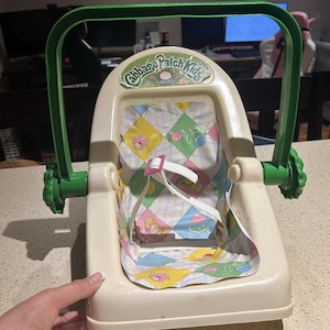 May include: A vintage Cabbage Patch Kids doll carrier featuring a cream-coloured plastic frame with a movable green handle. The seat insert has a white, yellow, green, and pink diamond-patterned fabric with small character illustrations and a white safety strap.