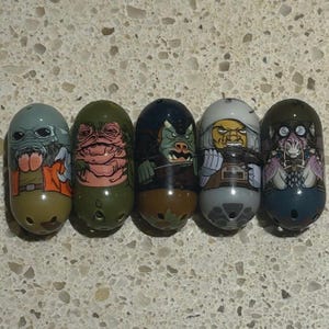 Può includere: Cinque giocattoli da collezione ovali e colorati con illustrazioni di personaggi. I giocattoli presentano vari disegni, tra cui una creatura con una testa grande e un gilet arancione e un mostro verde. I giocattoli sono su una superficie maculata.