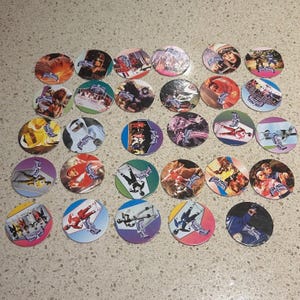 May include: A selection of vintage circular collectible game pieces, each with colourful illustrations of action figures and scenes. The pieces are arranged on a light surface, displaying a variety of designs and characters.