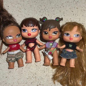 Puede incluir: Cuatro muñecas Bratz con diferentes peinados y atuendos. Las muñecas llevan ropa y accesorios coloridos, incluyendo una camisa roja y negra, una camisa morada con una rana verde, una camisa roja con un corazón rosa y una camisa negra con una mariposa.