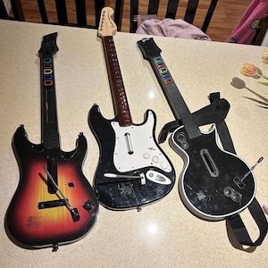May include: Three electric guitars, one red, one black, and one black with a white pickguard. The guitars are all for a video game and have coloured buttons on the fretboard.
