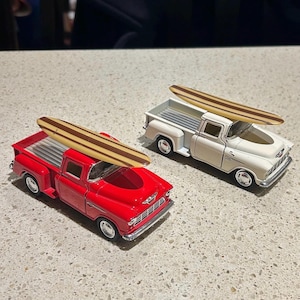 Puede incluir: Dos camionetas de juguete con tablas de surf en la parte superior. Una camioneta es roja y la otra es blanca.