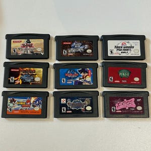 Könnte beinhalten: Neun schwarze Game Boy Advance-Cartridges werden gezeigt, mit Spieletiteln wie "Yu-Gi-Oh! The Sacred Cards", "Sonic Advance" und "Tiger Woods PGA Tour Golf". Die Cartridges haben farbenfrohe Spielgrafiken und Textetiketten.