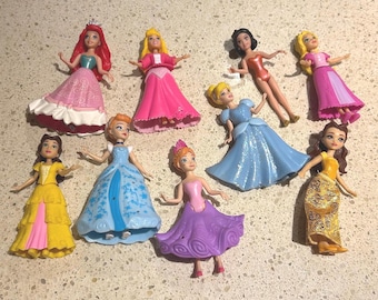 May include: A collection of nine small plastic princess figurines featuring various characters in colorful dresses, including red, pink, yellow, blue, and purple gowns. The dolls have painted facial features and molded hair, standing in different poses.