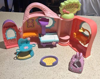 May include: A pink and purple toy playset with various accessories. The playset features a clock, a bed, a chair, and a pet carrier. The accessories are in shades of pink, purple, blue, and yellow. The playset is designed for imaginative play.