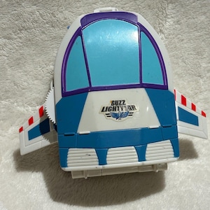 Può includere: Un'astronave giocattolo di Buzz Lightyear bianca e blu con dettagli viola e rossi. Il giocattolo ha un finestrino e ali con strisce rosse e bianche. Le parole "Buzz Lightyear" sono stampate sul davanti.