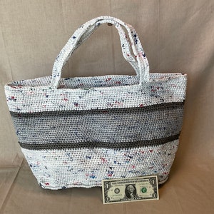 May include: White crocheted tote bag with a brown stripe and colorful flecks throughout. A one dollar bill is placed in front of the bag for scale.