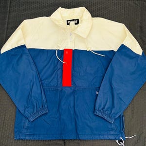 May include: A color-blocked anorak jacket in cream, navy blue, and red. The jacket features a half-zip closure, a stand-up collar, and a front pocket. The brand name "oest" is printed on the collar. The sleeves have elastic cuffs and drawstrings at the hem.