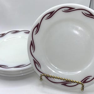 May include: A set of white plates with a maroon leaf-like design around the rim. The plates are stacked and displayed on a gold-colored plate stand. The plates have a glossy finish and are clean.