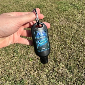 May include: A black liquid chalk bottle with a carabiner clip. The bottle has a blue and white label with the text "DOLPHSHOPDOG LIQUID CHALK" and "50ml". The bottle is held against a background of green grass.