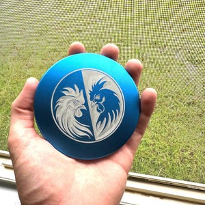Puede incluir: Un molinillo redondo de metal azul con un diseño plateado de dos gallos enfrentados. Los gallos están a ambos lados de una línea divisoria.