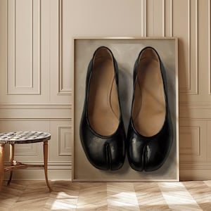 May include: A framed artwork depicting two black split-toe ballet flats. The shoes have a glossy finish and a neutral-toned interior. The artwork is set against a neutral backdrop, within a gold frame.