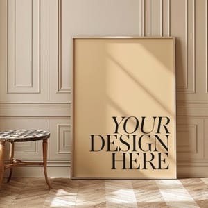 May include: A large framed poster with the text "YOUR DESIGN HERE" in black against a beige background. The poster is leaning against a wall with decorative molding. A small side table with a patterned top is in the foreground.