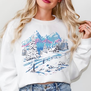 Pode incluir: Camisola branca com um design de paisagem de inverno. O gráfico retrata montanhas cobertas de neve, árvores perenes e uma pequena cabana. A paleta de cores inclui tons de azul, rosa e branco, criando uma serena cena de inverno. A camisola tem gola redonda e mangas compridas.
