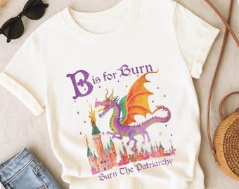 May include: Cream-coloured t-shirt with a dragon design. The dragon is purple, orange, and green, with the text "B is for Burn" and "Burn The Patriarchy". The design includes a castle and stars.