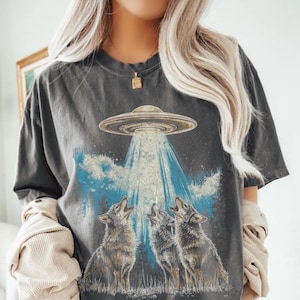 Puede incluir: Camiseta gris oscuro con un gráfico de un OVNI que emite luz sobre tres lobos aullando. El diseño incluye nubes azules y una silueta de bosque. La camiseta tiene un aspecto vintage y desgastado.
