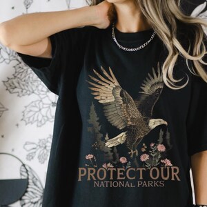Può includere: Maglietta nera con un'immagine di un'aquila marrone e dorata e il testo "PROTECT OUR NATIONAL PARKS".