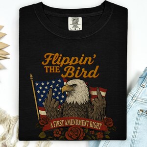 May include: Black t-shirt with a graphic of an eagle giving the middle finger in front of an American flag. The text reads "Flippin' the Bird" and "A First Amendment Right" in a vintage style.