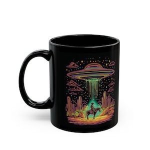 May include: Black ceramic mug featuring a colorful illustration of a UFO beaming a cowboy on horseback. The design includes desert cacti, stars, and a rainbow-colored UFO. The mug has a curved handle.