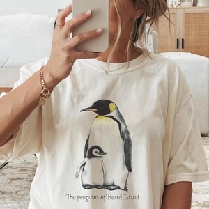 Könnte beinhalten: Weißes T-Shirt mit einem Aufdruck von zwei Pinguinen. Der größere Pinguin ist schwarz-weiß mit einem gelben Schnabel. Der kleinere Pinguin ist gelb-weiß. Der Text "The penguins of Heard Island" ist unter den Pinguinen aufgedruckt.