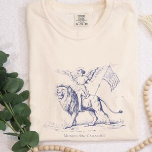 Könnte beinhalten: Ein cremefarbenes T-Shirt mit einem blauen Grafikdruck eines Engels, der auf einem Löwen reitet. Der Engel hält eine Flagge mit Sternen und Streifen. Der Text "MUNDUS SINE CAESARIBUS" ist unter dem Grafikdruck gedruckt.