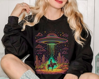 May include: A black sweatshirt with a colorful graphic of a UFO hovering over a cowboy riding a horse in a desert landscape. The graphic is outlined in neon colors and includes stars and clouds.