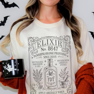 以下が含まれることがあります： 「ELIXIR No. 8647」とボトルイラストが描かれた、ヴィンテージスタイルのグラフィックが特徴のクリーム色のTシャツ。「Banishing Orange Phantoms Since 2016」の文字も含まれています。魔女のシルエットが描かれた黒いマグカップが画像に写っています。