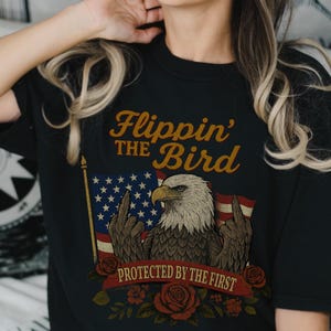 Può includere: T-shirt nera con una grafica di un'aquila calva americana che mostra il dito medio davanti a una bandiera americana. Il testo "Flippin' the Bird" e "Protected by the First" è presente.