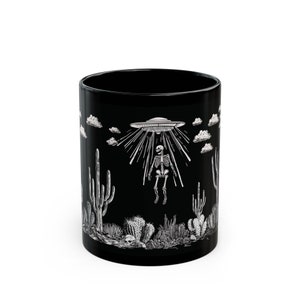 May include: Black ceramic mug featuring a white graphic of a skeleton being abducted by a UFO in a desert landscape with cacti.
