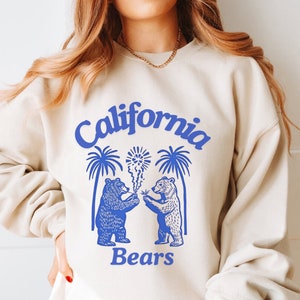 May include: A cream-colored sweatshirt with a blue graphic design featuring two bears holding sparklers under palm trees. The text "California Bears" is printed below the bears.