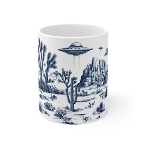Könnte beinhalten: Weiße Keramik-Tasse mit einer blau-weißen Illustration einer Wüstenlandschaft mit Kakteen, Felsformationen und fliegenden Untertassen. Das Design hat eine Vintage-Ästhetik im Toile-Stil.