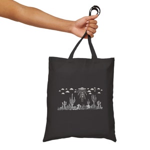 May include: Black tote bag with a white graphic of a skeleton being abducted by a UFO in a desert landscape with cacti.