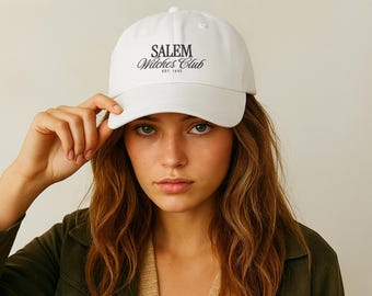 May include: A white baseball cap with the text "SALEM Witches Club EST. 1692" in black lettering. The cap is being held by a person.