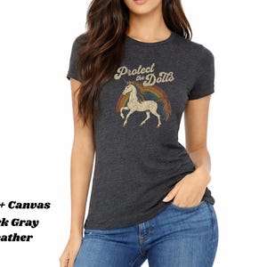 May include: Dark gray heather t-shirt with a vintage-style graphic. The graphic features a unicorn, a rainbow, and the text "Protect the Dolls" in a retro font. The shirt is made of Bella + Canvas material.