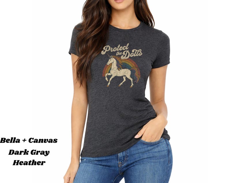 May include: Dark gray heather t-shirt with a vintage-style graphic. The graphic features a unicorn, a rainbow, and the text "Protect the Dolls" in a retro font. The shirt is made of Bella + Canvas material.