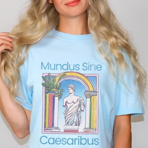 Könnte beinhalten: Hellblaues T-Shirt mit einem grafischen Druck einer Statue in einem bunten Torbogen. Der Text "Mundus Sine Caesaribus" ist auf dem Shirt aufgedruckt.